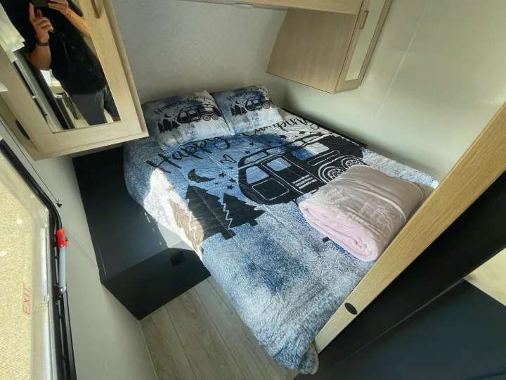 Inside an RV rental: a neat bedroom with "Happy Camping" bedspread, pink blanket, and modern light wood decor.. Prime Time Avenger 2024