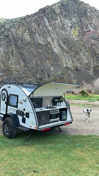 Teardrop RV rental with an open outdoor kitchen parked by rocky mountains. A Dalmatian dog walks on grass. Ideal for camping.. nuCamp T@G 2023