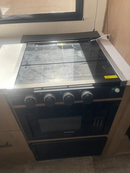 This RV rental kitchen features a GREYSTONE stove with a glass cover (raise before cooking) and a FURRION microwave oven below.. Forest River Campsite reserve 27MR 2025