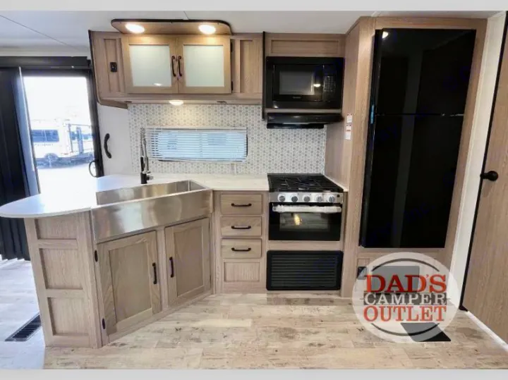 This modern RV rental kitchen features a stainless steel farmhouse sink, light wood cabinets, gas range, microwave, and black fridge.. Gulf Stream Innsbruck 2024