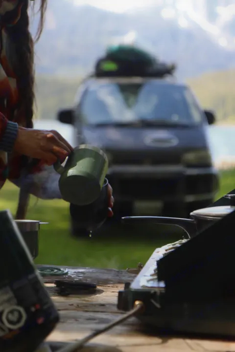 A person pours steaming coffee outdoors from a green mug. Camping stove & dark van (RV rental) visible by a lake & mountains.. Mitsubishi Delica 2000