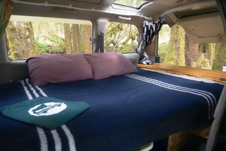 An RV rental's cozy bed with a dark blue blanket, pillows, and green logo bag, offering serene forest views.. MITSUBISHI Delica 1997