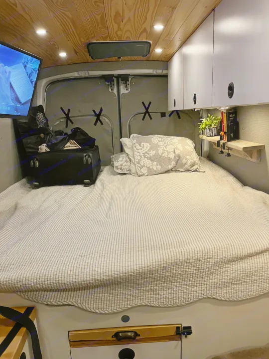 Cozy RV rental interior shows a comfortable bed, TV, overhead cabinets, and wooden ceiling in a camper van.. Ford Custom Transit 2019