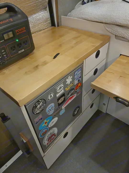 Inside an RV rental: A Jackery power station sits on a light wood counter with a sticker-covered fridge and storage drawers below.. Ford Custom Transit 2019