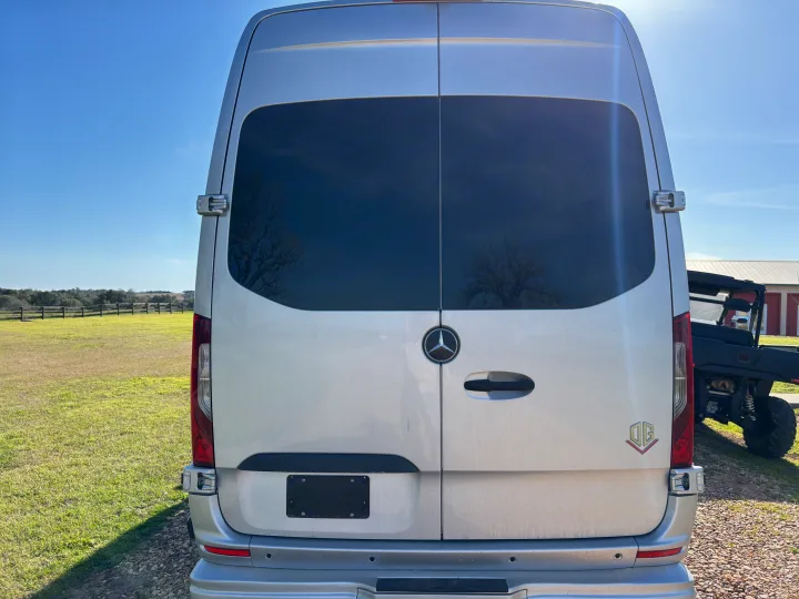 Rear view of a silver Mercedes-Benz Sprinter **RV rental** van, with dark rear windows, parked in a sunny, grassy field.. Mercedes-Benz OG Luxury Coach V Cruis 2025