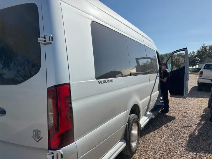 Silver RV rental van parked on gravel, with a man entering through the open side door.. Mercedes-Benz OG Luxury Coach V Cruis 2025