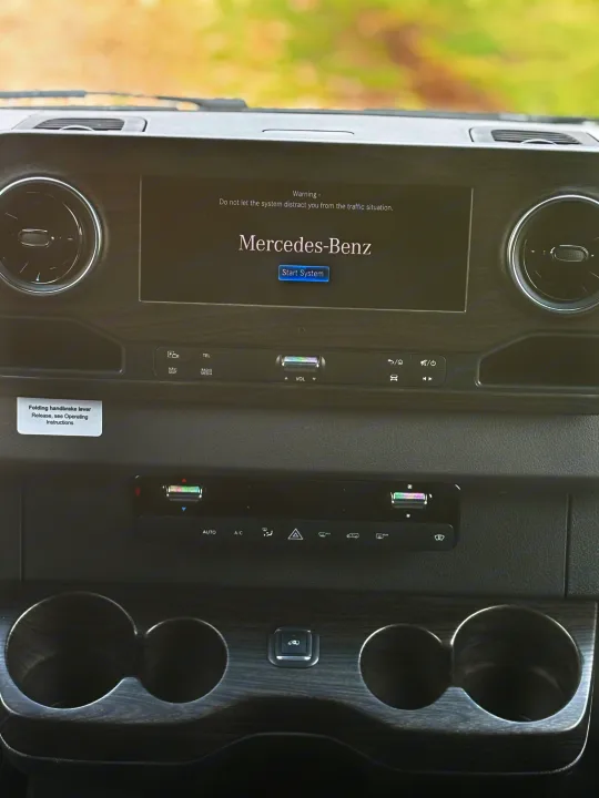 Inside a Mercedes-Benz RV rental: the dashboard features an infotainment screen with a warning, climate controls, and cup holders.. Mercedes-Benz OG Luxury Coach V Cruis 2025