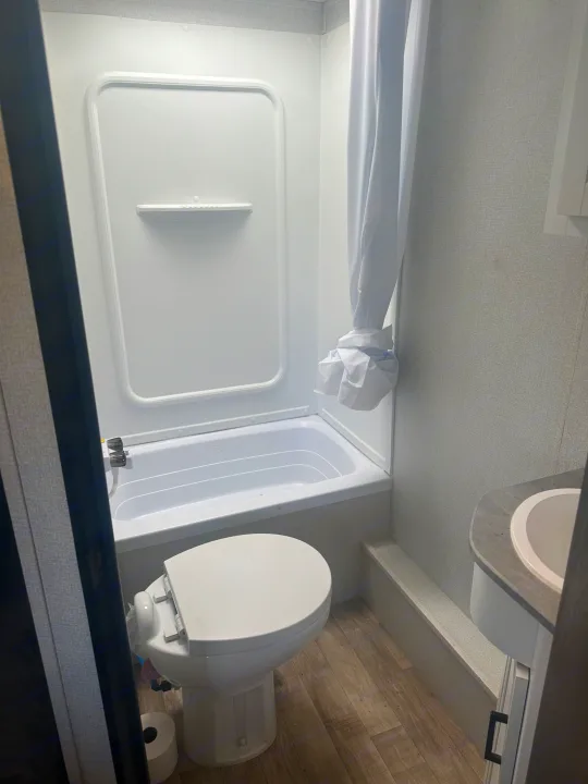 Compact RV rental bathroom features a white toilet, small tub/shower, and sink vanity. Light walls and wood-look floor.. Keystone Hideout 2022