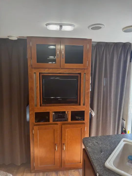 Inside an RV rental: a wooden entertainment unit with TV & stereo, cozy curtains, and a kitchen sink area.. Keystone RV Passport Grand Touring 2014