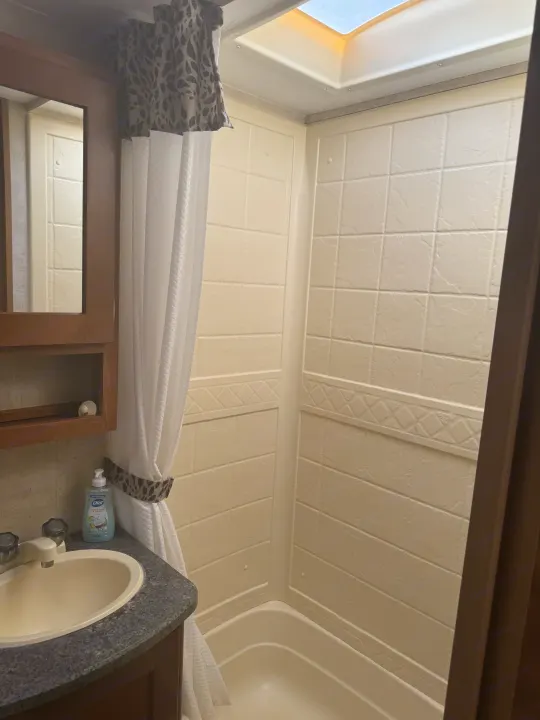 An RV rental bathroom with a beige tiled-look shower stall, a leopard-print curtain, a sink, and a skylight.. Keystone RV Passport Grand Touring 2014