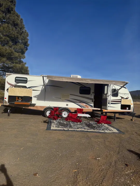 A white travel trailer RV rental at a sunny campsite, equipped with an outdoor kitchen, awning, table, and chairs.. Keystone RV Passport Grand Touring 2014