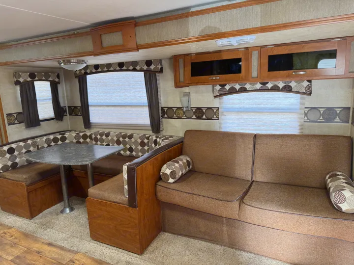 Inside an RV rental, a cozy living area with a brown fabric sofa, a dinette booth with patterned cushions, overhead cabinets, and large windows.. Keystone RV Passport Grand Touring 2014