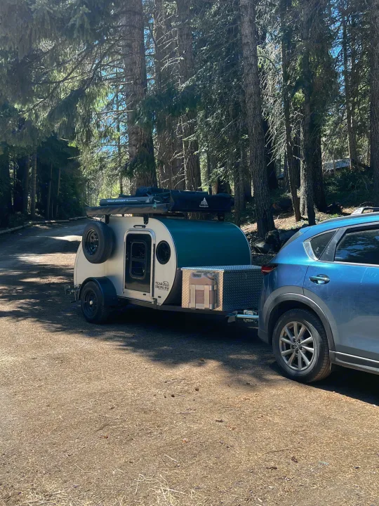 An RV rental teardrop camper hitched to a blue SUV on a sunny forest road. Perfect for camping adventures.. Teardrops Northwest Trek 2022