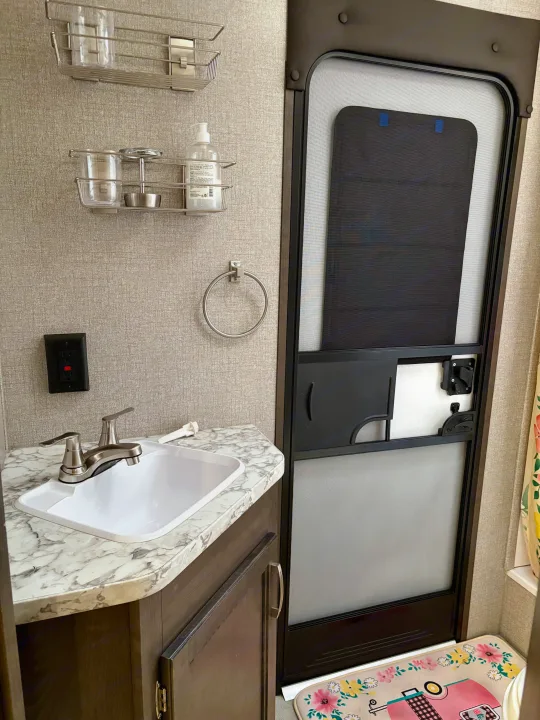 An RV rental bathroom, featuring a compact sink, faucet, countertop, wall storage, and interior door with screen and shade.. Jayco Jay Flight SLX 2021