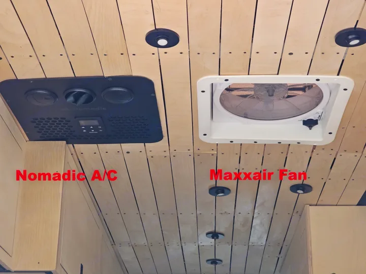 Ceiling view inside an RV rental, showing a black Nomadic A/C unit on the left and a white Maxxair Fan on the right, on light wood panels.. mercedes 144 High Roof AWD 2024