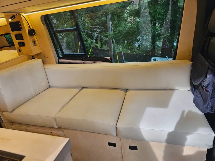 Inside an RV rental: light wood interior with a long beige bench seat, storage, and a large window showcasing outside trees.. mercedes 144 High Roof AWD 2024