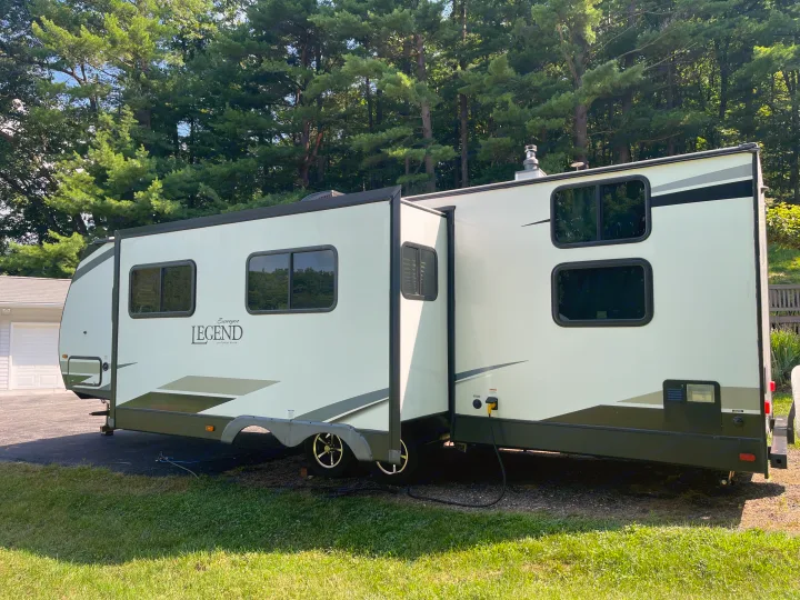 A large, modern Surveyor Legend RV rental, light-colored with dark accents, is parked and set up for use among green trees.. Forest River Surveyor Legend 2019