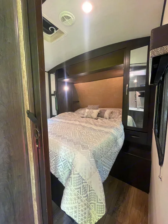 Inviting RV rental bedroom features a patterned queen bed, dark wood built-ins, mirror, and ceiling lights.. Forest River Surveyor Legend 2019