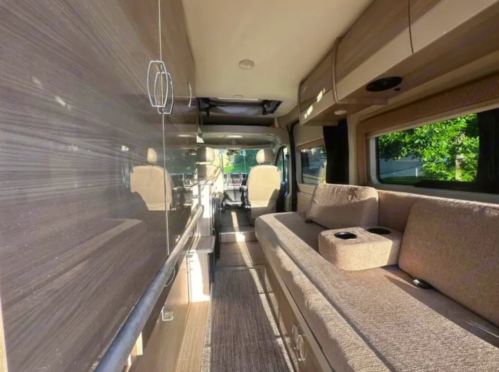 Inside this modern RV rental: a cozy living space with a long couch, overhead cabinets, and bright views.. Thor Motor Coach 18T 2022