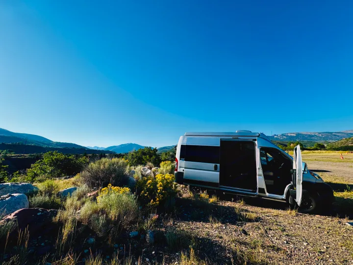 A silver camper van (RV rental) is parked with open doors in a scenic, mountainous landscape under a blue sky.. Thor Motor Coach 18T 2022