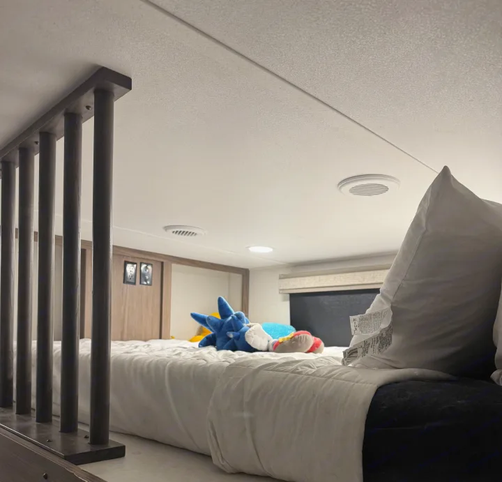 Inside an RV rental: a comfortable loft bed with a white comforter, Sonic plushie, dark wood railing, and ceiling vents.. Alliance Paradigm 2023