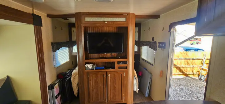 RV rental interior. A central wood entertainment unit with a TV separates spaces, featuring windows, blinds, storage, and an open door to outside.. Heartland RVs Prowler Lynx 2018
