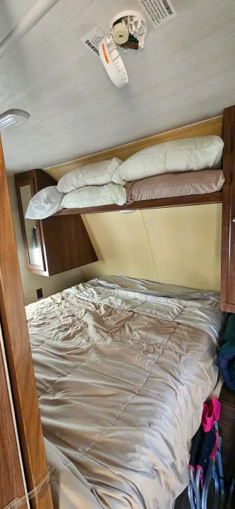 This RV rental bedroom features a bed with comforter, pillows on an overhead shelf, and a disassembled smoke detector on the ceiling.. Heartland RVs Prowler Lynx 2018