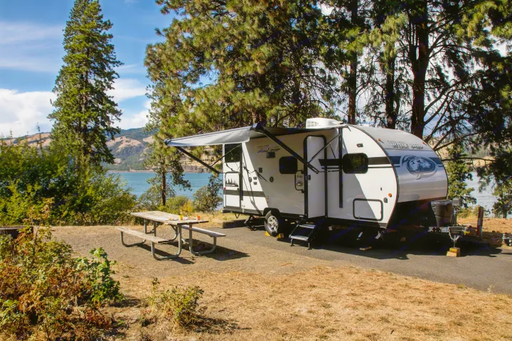 Scenic rv rental setup: a white travel trailer with awning deployed, parked at a lakeside campsite with a picnic table, under tall trees.. Forest River Cherokee Wolf Pup Black Label 2020