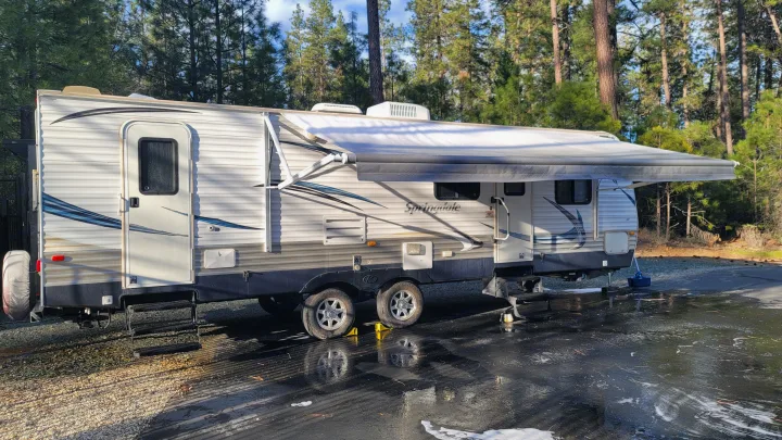 RV rental travel trailer with awning extended, parked on wet pavement in a wooded area, likely being cleaned.. Keystone RV Springdale 2012