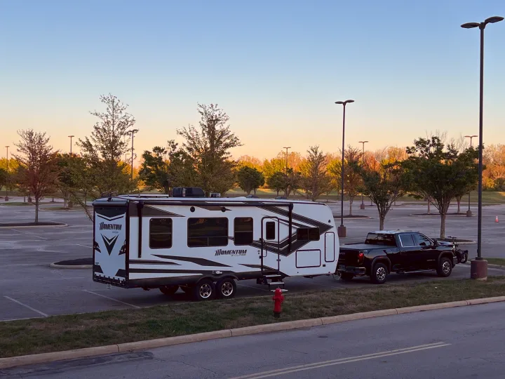 Black truck towing a white "Momentum" travel trailer in a large parking lot, perfect for an rv rental journey.. Grand Design Momentum Toy Hauler 2022