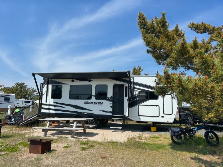A white Momentum RV rental (travel trailer) set up at a sunny campsite with an e-bike and picnic table.. Grand Design Momentum Toy Hauler 2022