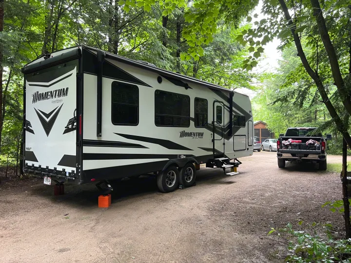 A white Grand Design Momentum RV rental, a toy hauler, is perfectly set up at a shaded forest campsite with a pickup truck.. Grand Design Momentum Toy Hauler 2022