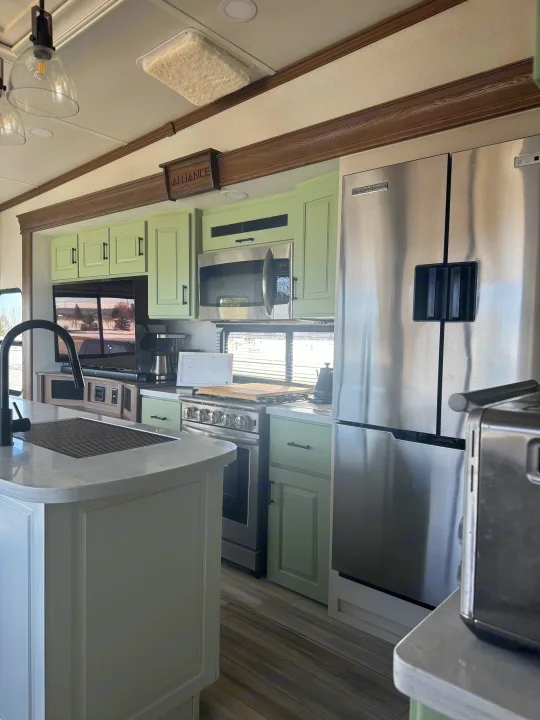 Modern RV rental kitchen with light green cabinets, stainless steel refrigerator, range, microwave, and a central island.. Alliance Paradigm 2023