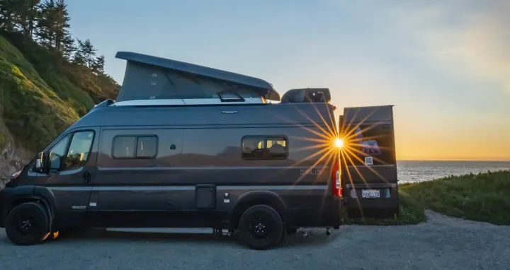 A dark grey RV rental with its pop-top extended, parked on a coastal bluff, enjoying a vibrant sunset over the ocean.. Winnebago Solis 59PX 2023