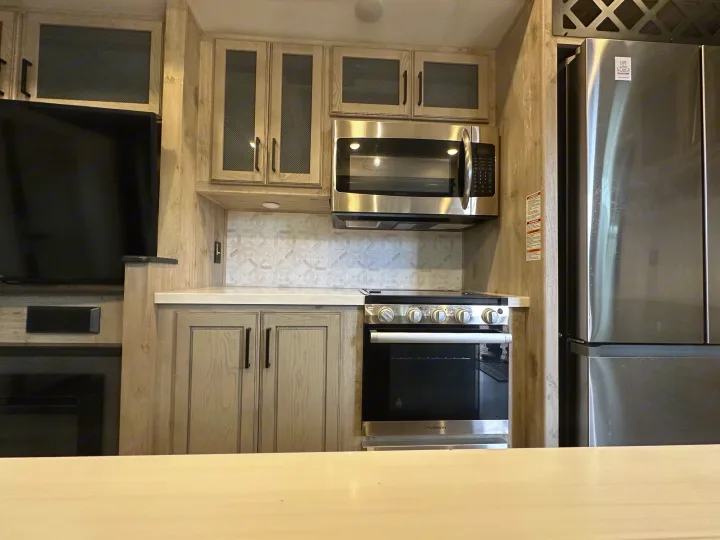 Well-appointed RV rental kitchen showing stainless fridge, microwave, oven, light wood cabinets, and patterned backsplash.. Keystone RV Avalanche 2023