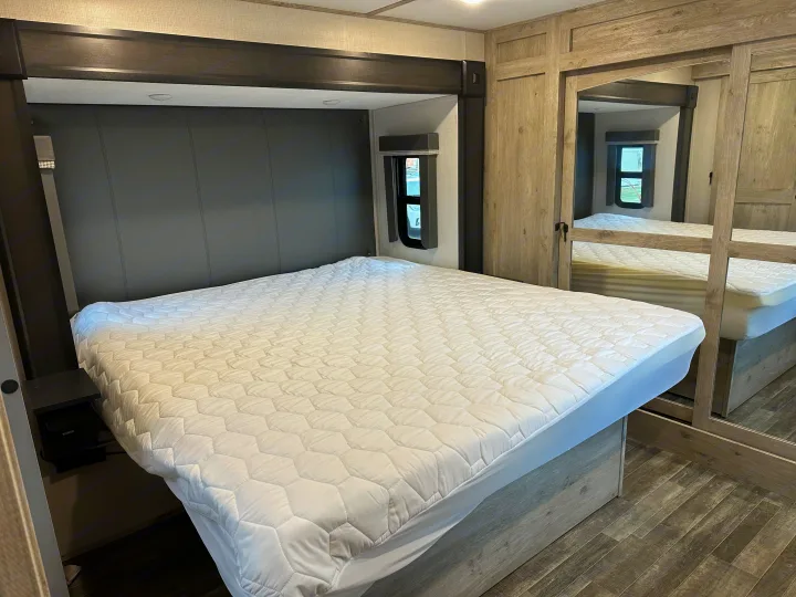 Comfortable RV rental bedroom featuring a large bed with a quilted mattress pad, window, nightstand, and mirrored closet.. Keystone RV Avalanche 2023