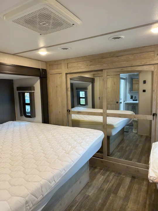 Inside an RV rental: Master bedroom with mirrored wardrobe reflecting bathroom, plush bed, and modern wood-style floor.. Keystone RV Avalanche 2023