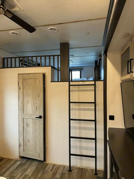 Inside an RV rental: a modern loft sleeping area with a ladder, a light-colored door, and a ceiling fan overhead.. Keystone RV Avalanche 2023