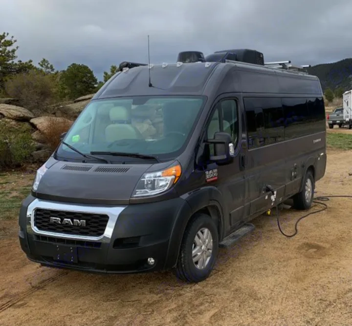 Dark grey Ram ProMaster RV rental parked on dirt, hooked up. Mountains & trees under cloudy sky. Ideal for camping.. Thor Sequence 2021