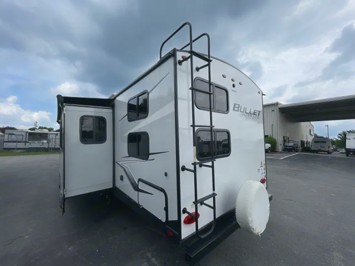 A white Bullet Ultra Lite travel trailer (RV) with an extended side, rear ladder, and spare tire, ready for an rv rental.. Keystone Bullet 2023