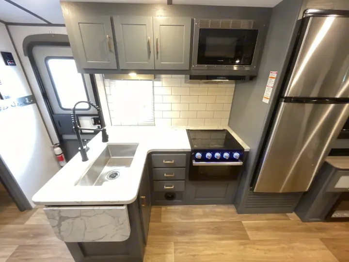A modern RV rental kitchen featuring a stainless steel sink, stove, microwave, refrigerator, gray cabinets, and white subway tile backsplash.. Keystone Bullet 2023