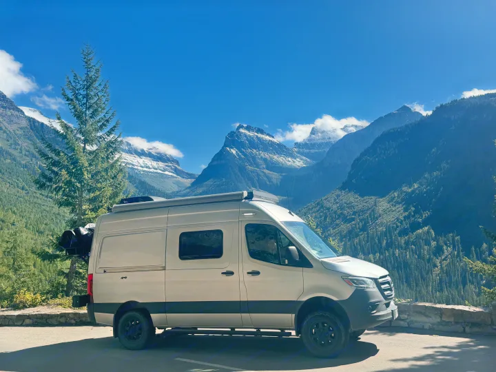Explore nature's beauty! A light-colored RV rental is parked at a scenic mountain overlook, surrounded by snow-capped peaks and tall trees.. Winnebago Revel 2021