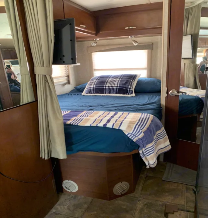 Compact RV rental bedroom with a bed, blue bedding, plaid pillow, striped blanket, TV, and window.. Fleetwood Tioga 2011