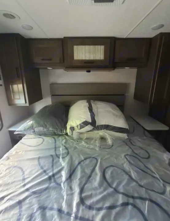 RV rental bedroom interior: A cozy bed with new bedding, wrapped in plastic, surrounded by compact cabinets for storage.. Coachmen Mirada 2024