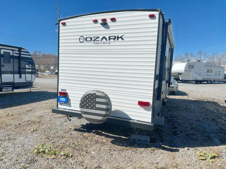 Rear view of an Ozark Forest River travel trailer (RV rental) on a gravel lot. Features a grayscale American flag tire cover.. Ozark Lite 2021