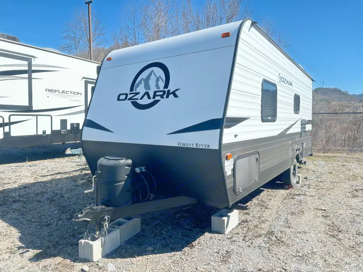 White & dark grey Ozark travel trailer, likely an RV rental, next to a Reflection RV on a sunny lot.. Ozark Lite 2021