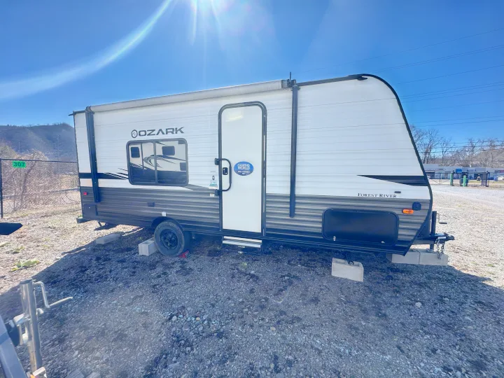 A white and gray pre-owned Forest River Ozark travel trailer for an RV rental, parked on gravel under a sunny sky.. Ozark Lite 2021