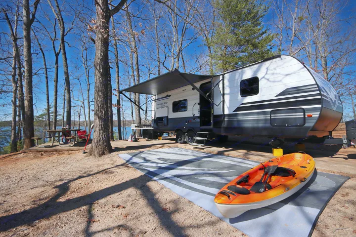 Lakeside RV rental campsite features a travel trailer with an awning, kayak, and picnic area, ideal for nature lovers.. Grand Design Transcend Xplor 2020