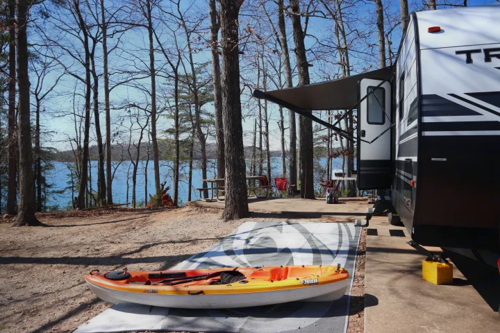 A scenic lakeside RV rental campsite, featuring an extended awning, kayak, and picnic spot under bare trees.. Grand Design Transcend Xplor 2020