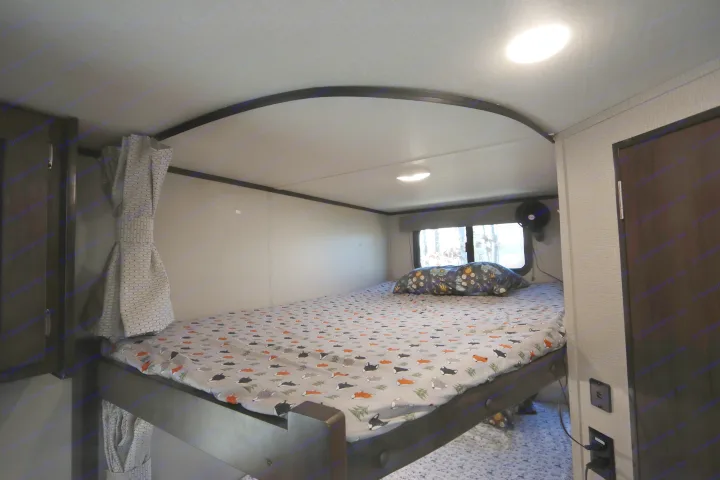 Cozy bunk bed sleeping area inside an RV rental, complete with a patterned sheet, window view of trees, and personal fan.. Grand Design Transcend Xplor 2020